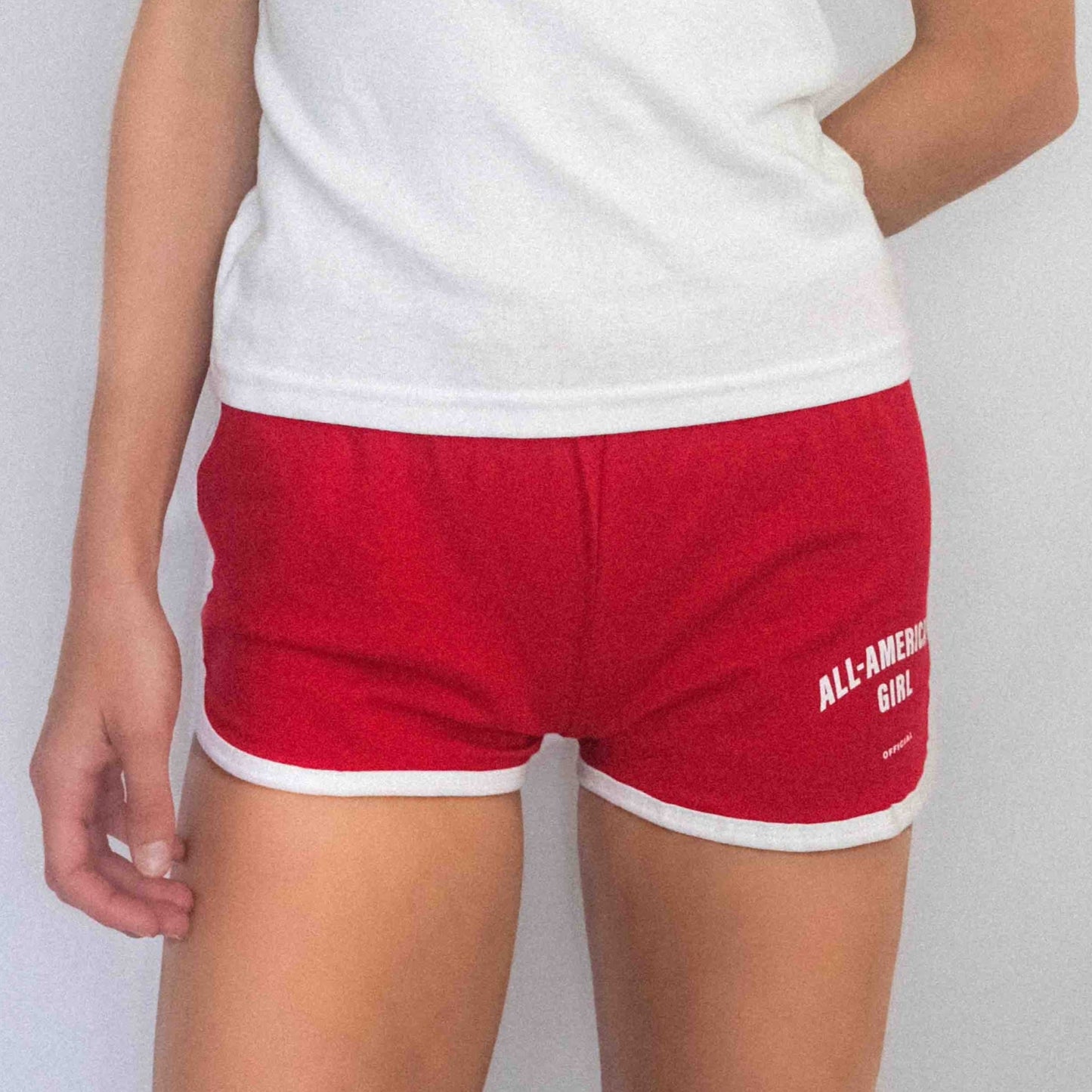 A girl wearing red All-American Girl track shorts. The shorts say All-American Girl Official on them. This is a comfortable pair of shorts perfect for working out, lounging, or casual style. These shorts come in small, medium, and large.