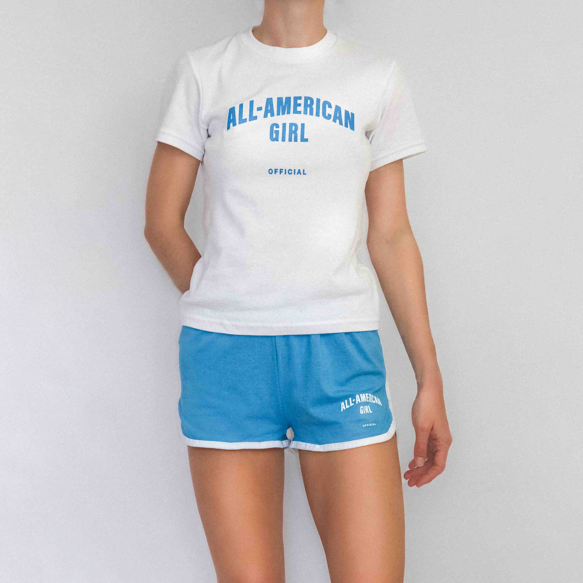 A girl wearing a blue All-American Girl baby tee. The tee says All-American Girl Official on it. This is a comfortable, fitted white tee with blue text. This tee is one size.