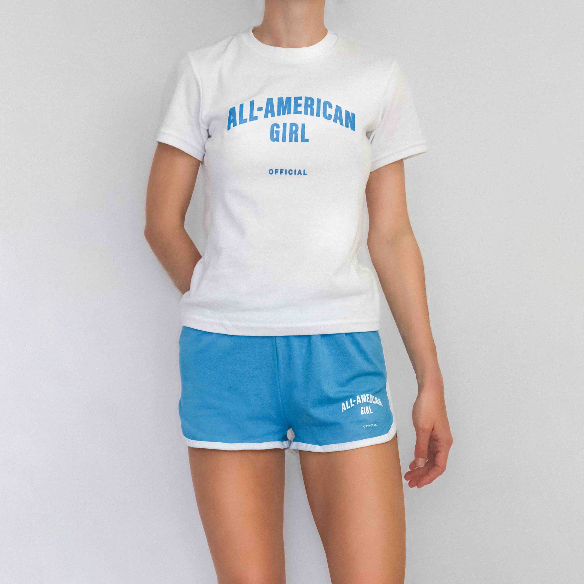 A girl wearing blue All-American Girl track shorts. The shorts say All-American Girl Official on them. This is a comfortable pair of shorts perfect for working out, lounging, or casual style. These shorts come in small, medium, and large. The shorts are paired with the matching tee.