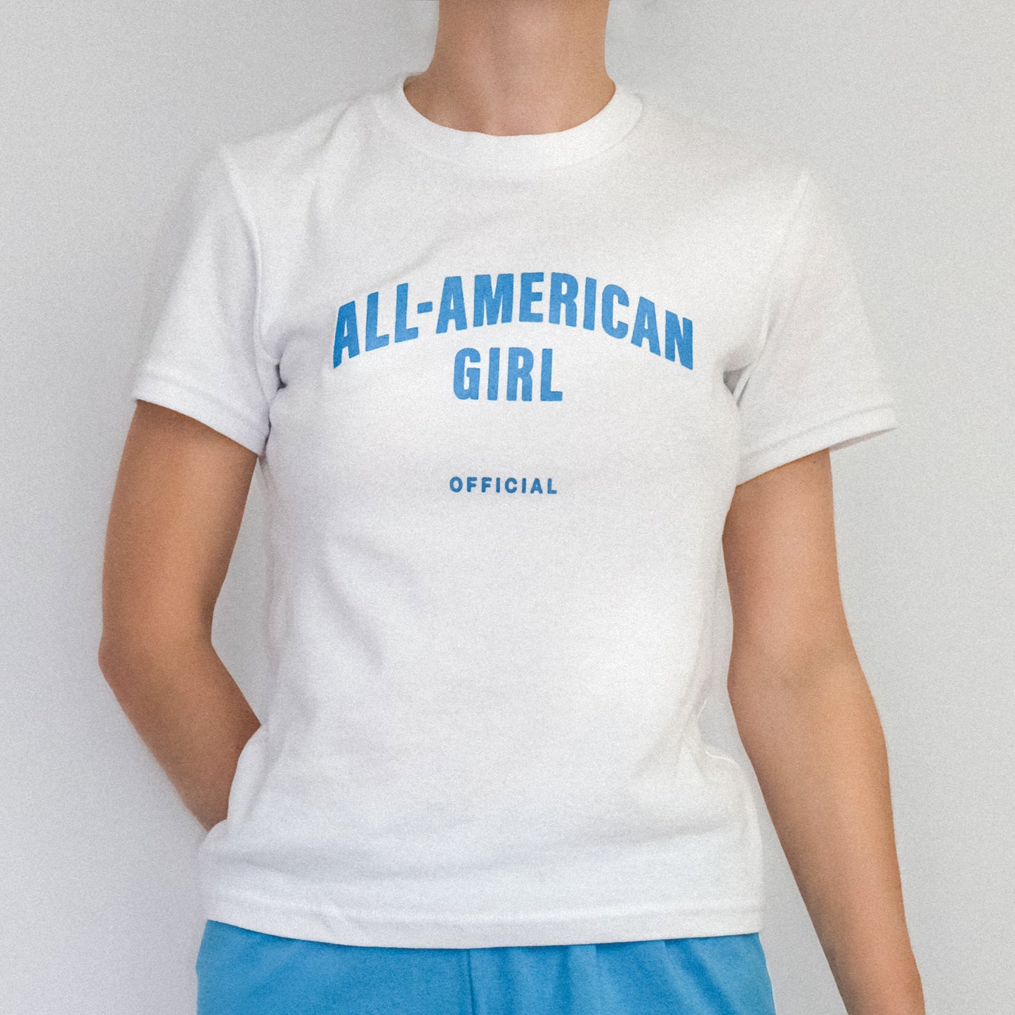 A girl wearing a blue All-American Girl baby tee. The tee says All-American Girl Official on it. This is a comfortable, fitted white tee with blue text.