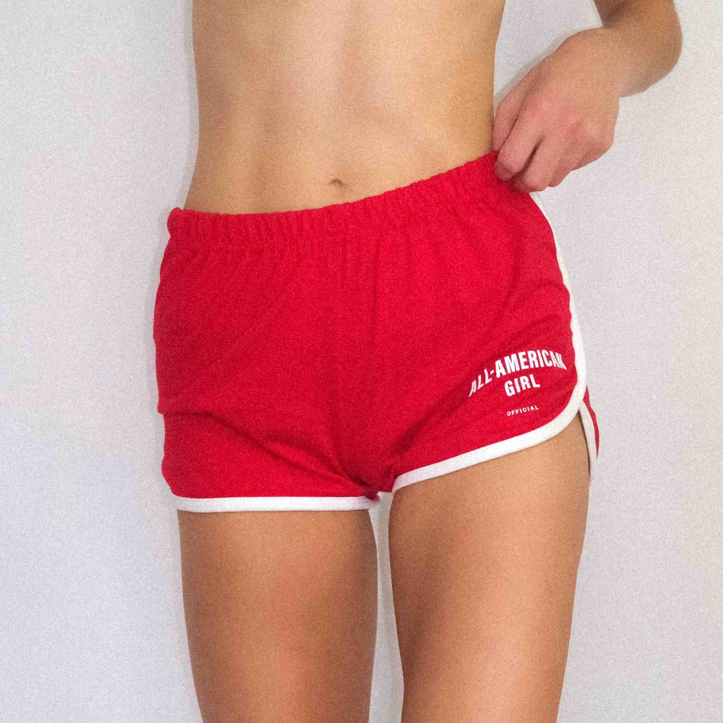 A girl wearing red All-American Girl track shorts. The shorts say All-American Girl Official on them. This is a comfortable pair of shorts perfect for working out, lounging, or casual style. These shorts come in small, medium, and large.